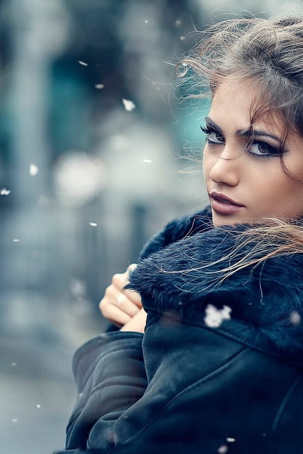 Winter hairstyles — woman in a cozy winter coat with wind-swept hair and falling snow, front portrait